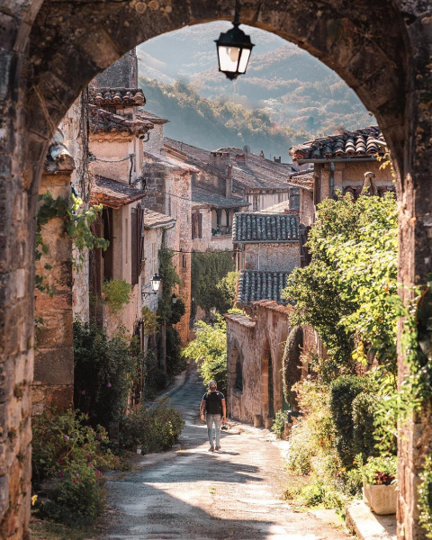 Photo d’une ruelle de village ancien pittoresque avec maisons en pierre, végétation et montagnes au fond.