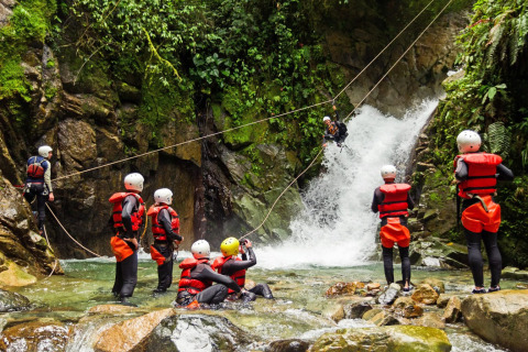 Group of people in life jackets and helmets enjoying ziplining and canyoning near a waterfall at a holiday park.