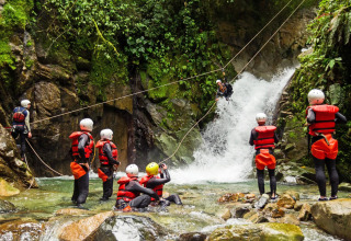 Gruppo di persone con giubbotti di salvataggio e caschi fa zipline e canyoning presso una cascata in un parco.
