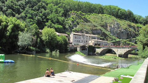 Foto del campeggio Camp Redon in Occitania, Francia, con fiume, ponte in pietra, case e colline verdi.