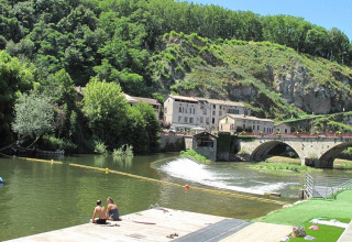 Foto del campeggio Camp Redon in Occitania, Francia, con fiume, ponte in pietra, case e colline verdi.