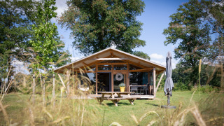 Foto der Wellness Luxe Lodge, eine gemütliche Holzhütte mit Terrasse im Grünen, umgeben von Bäumen.