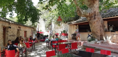 Area caffè all’aperto con sedie rosse e ospiti rilassati sotto alberi ombrosi al campeggio Le Moulin de Cost.