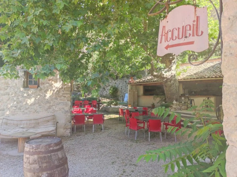 Zona esterna del campeggio Le Moulin de Cost in Francia con sedie rosse sotto alberi ombrosi.