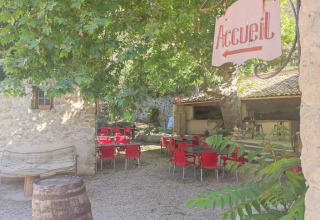 Zona esterna del campeggio Le Moulin de Cost in Francia con sedie rosse sotto alberi ombrosi.