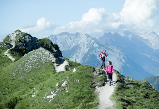 Twee wandelaars in roze jassen lopen op een bergpad bij Pettneu am Arlberg met Alpen op de achtergrond.