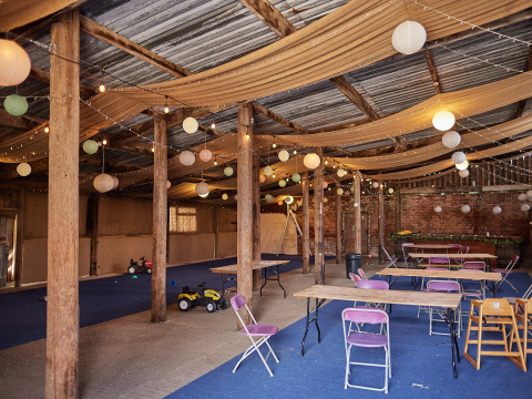 Intérieur d'une grange décorée pour un événement avec tables en bois, chaises pliantes et lanternes suspendues.