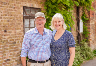 Una pareja mayor sonriente delante de un edificio de ladrillo en BoerenBed Moor Farm, suroeste de Inglaterra.