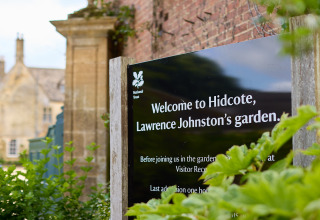 Bienvenido a Hidcote, el jardín de Lawrence Johnston, cartel frente a edificio de ladrillo y plantas en West Midlands, Reino Unido.