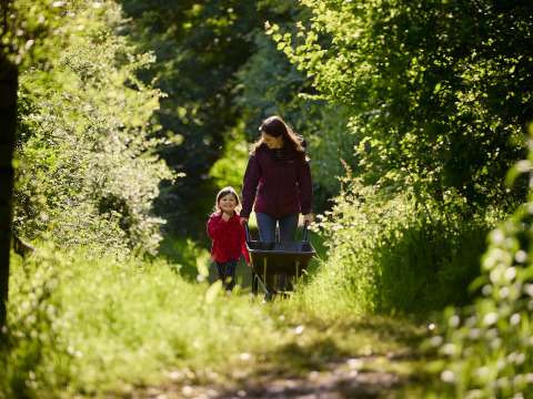 Kvinde og barn går sammen og skubber en trillebør på en solrig sti i BoerenBed Hidcote Manor Farm.
