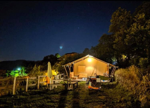 Ein beleuchtetes Safarizelt bei Nacht, umgeben von Bäumen, Bänken und einer natürlichen Landschaft.