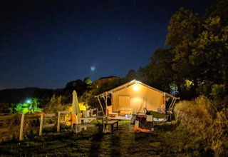 A safari tent illuminated at night, surrounded by trees, benches, and a rustic landscape outdoors.