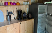 Pequeña cocina en una tienda safari con tazas, cafetera, hervidor eléctrico y nevera abierta.