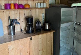 Petite cuisine dans une tente safari avec tasses, cafetière, bouilloire électrique et réfrigérateur ouvert.