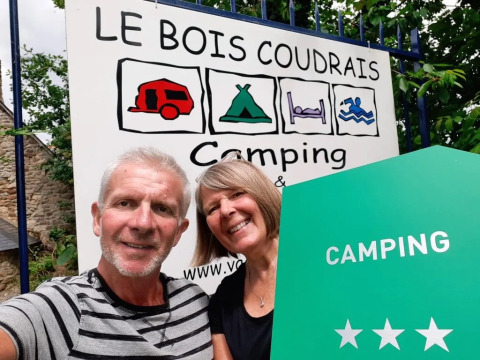 Coppia sorridente davanti all’insegna del campeggio Le Bois Coudrais in Bretagna, Francia, con tre stelle.