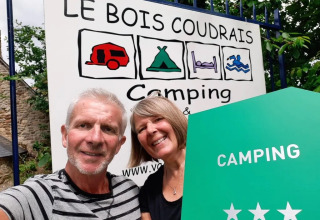 Coppia sorridente davanti all’insegna del campeggio Le Bois Coudrais in Bretagna, Francia, con tre stelle.