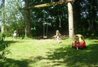 Bambini che giocano su un prato con altalene e una macchinina sotto gli alberi al campeggio Le Bois Coudrais in Bretagna.