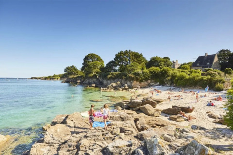 Spiaggia soleggiata con rocce, persone e una casa tra il verde vicino a Cuguen, Bretagna, Francia.