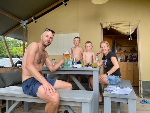 Eine Familie sitzt lachend an einem Picknicktisch vor einem Safarizelt und genießt eine Mahlzeit.