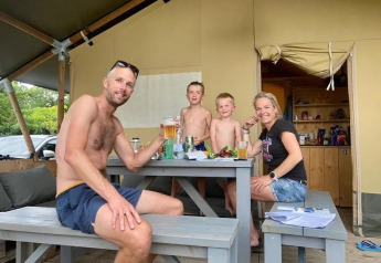 A family smiles while enjoying a meal and drinks together outside their safari tent at a campsite.