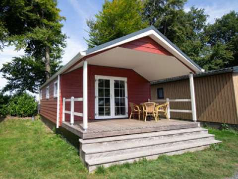 Luxe chalet rosso con veranda coperta, tavolo e sedie in vimini, circondato dal verde.