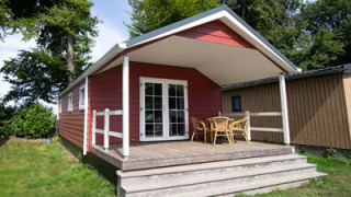 Luxe chalet rosso con veranda coperta, tavolo e sedie in vimini, circondato dal verde.