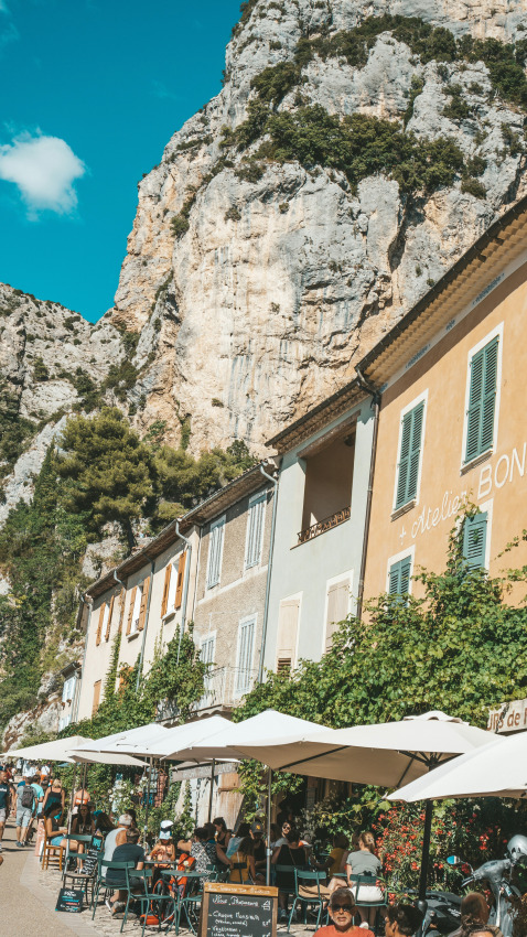Terrasse de café animée sous des parasols, au pied de maisons colorées et de falaises en Provence-Alpes-Côte d’Azur.