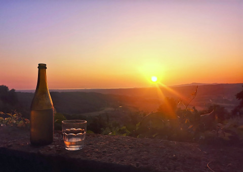 Een fles en een glas op een stenen muurtje bij zonsondergang met zicht op de heuvels rond Montescudaio, Toscane.