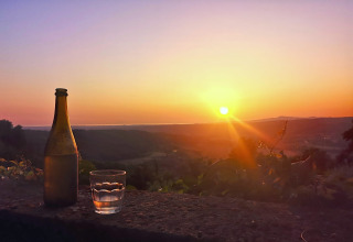 Een fles en een glas op een stenen muurtje bij zonsondergang met zicht op de heuvels rond Montescudaio, Toscane.