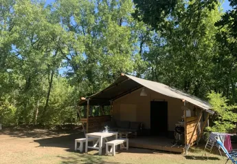 Tenda safari con veranda e tavolo all'aperto, immersa in una radura soleggiata tra alberi verdi.