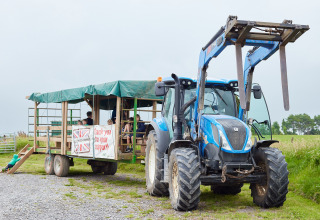 Blauwe tractor met overdekte aanhanger en bezoekers op BoerenBed Warren Farm vakantiepark in Zuidwest-Engeland.