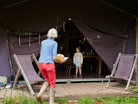 Woman in red pants and boots brings bread to a child outside a tent at BoerenBed Upper Shadymoor Farm, Wales.