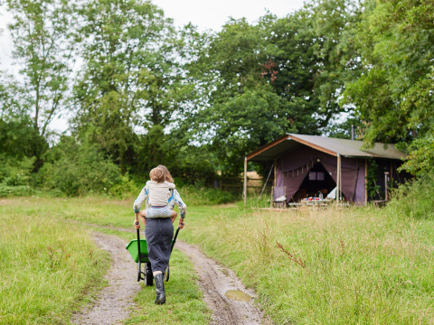 Adulto con bambino in spalla spinge una carriola su un sentiero erboso verso una tenda da glamping.