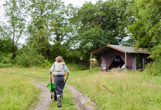Volwassene met kind in draagzak duwt een kruiwagen over een pad richting safaritent op een groene weide.
