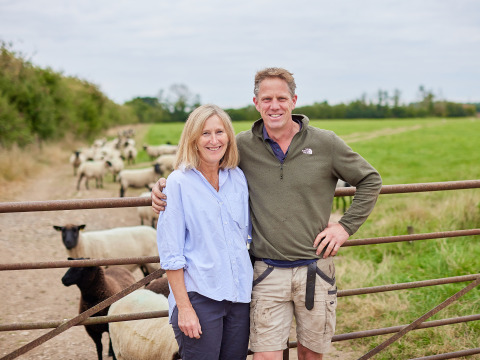 Zwei Menschen posieren lächelnd vor einem Weidezaun mit Schafen auf BoerenBed New Barn Farm in Ostengland.