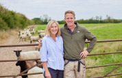 Couple souriant devant une barrière de ferme avec des moutons à BoerenBed New Barn Farm, Est de l’Angleterre.