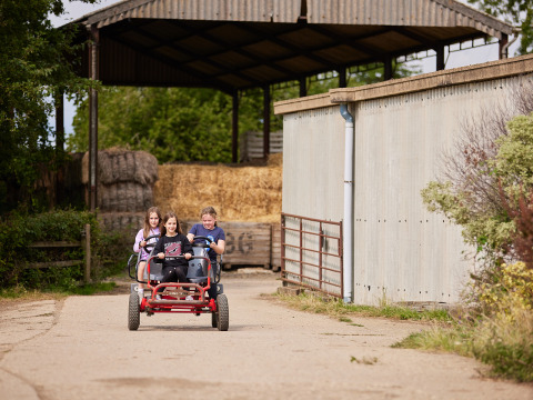 Tre børn cykler på en firehjulet gokart ved et landbrug med halmballer på BoerenBed New Barn Farm.