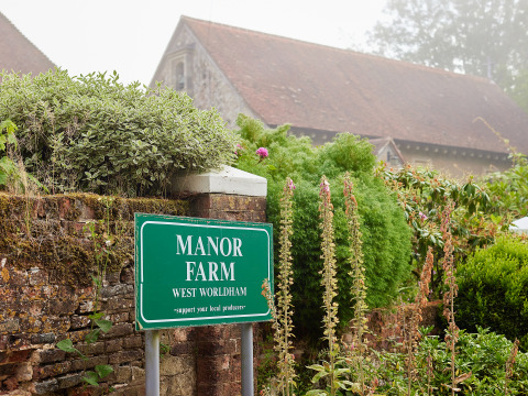 Cartello verde di Manor Farm a West Worldham, circondato da piante rigogliose e muro di mattoni in Inghilterra.