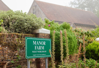Grünes Schild von Manor Farm in West Worldham, umgeben von üppigem Grün und Ziegelmauer, in Südostengland.