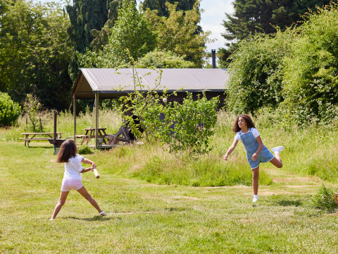 Two girls play rounders on a grassy field in front of a tent at BoerenBed Manor Farm holiday park, South East England.