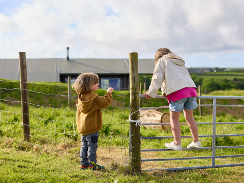 Dos niños juegan cerca de una valla en una granja con campos verdes y un granero al fondo en el suroeste de Inglaterra.