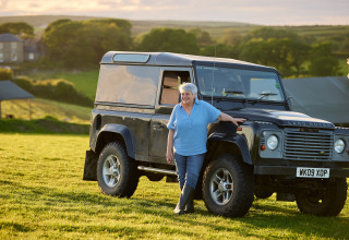 Eine ältere Frau steht lächelnd neben einem Land Rover auf einer Wiese der Treganhoe Farm im Südwesten Englands.