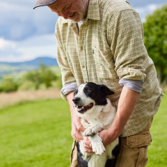 Man in geruite hemd speelt met zwart-witte hond op het gras aan BoerenBed Pant y March Farm, Wales.