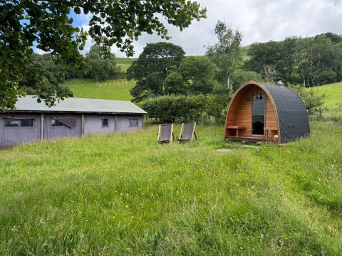 Træhytte og liggestole på en grøn eng ved BoerenBed Pant y March Farm i Wales, omgivet af bakker og træer.