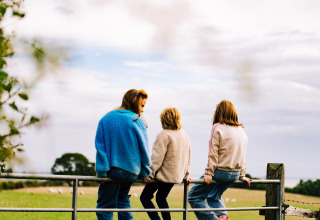 Three people sit on a fence overlooking fields at BoerenBed Lunsford Farm holiday park in South East England, UK.