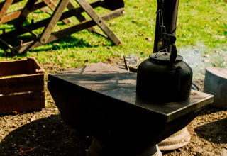A black kettle sits on an outdoor stove at BoerenBed Sunninglye Farm, with grassy surroundings and morning light.