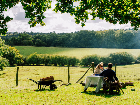 Ein Paar genießt ein Picknick an einem Tisch auf einer Wiese mit Blick auf Felder bei Sunninglye Farm, England.