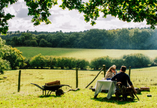 Ein Paar genießt ein Picknick an einem Tisch auf einer Wiese mit Blick auf Felder bei Sunninglye Farm, England.