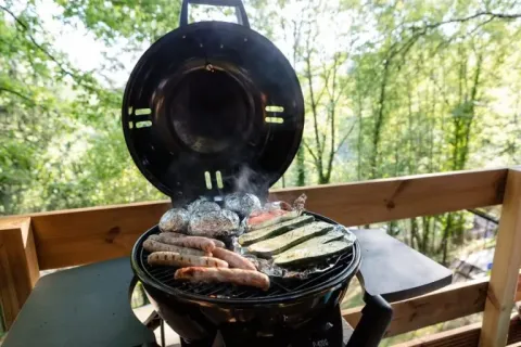 Cibo alla griglia su barbecue in veranda di tenda safari con vista sulla foresta, salsicce e verdure visibili.