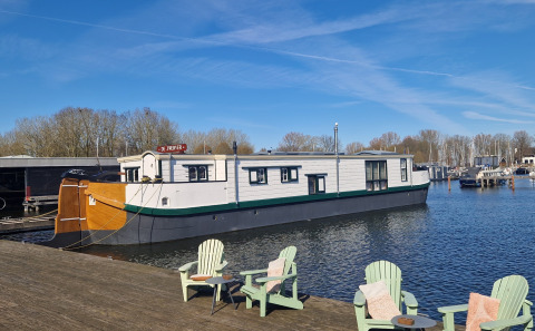 Péniche De Zwerver amarrée à un quai en bois avec des chaises vertes sous un ciel bleu clair.